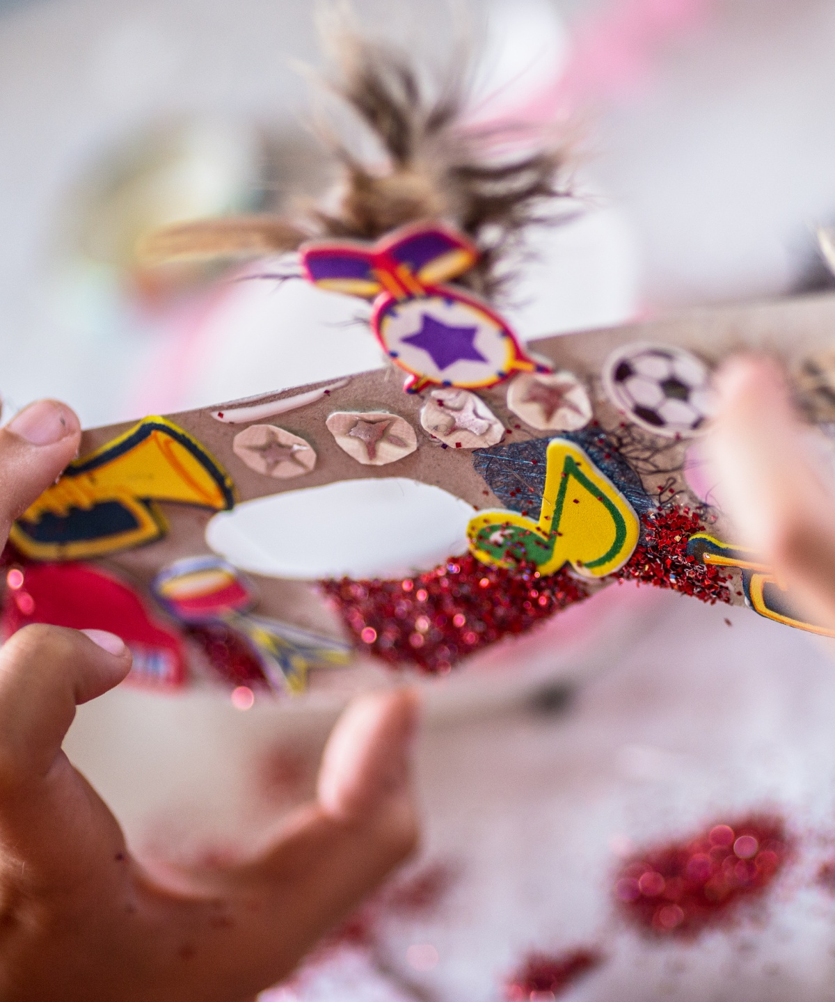 Person hält eine Maske mit Glitzer, Stickern und Federn in den Händen auf einem Basteltisch.