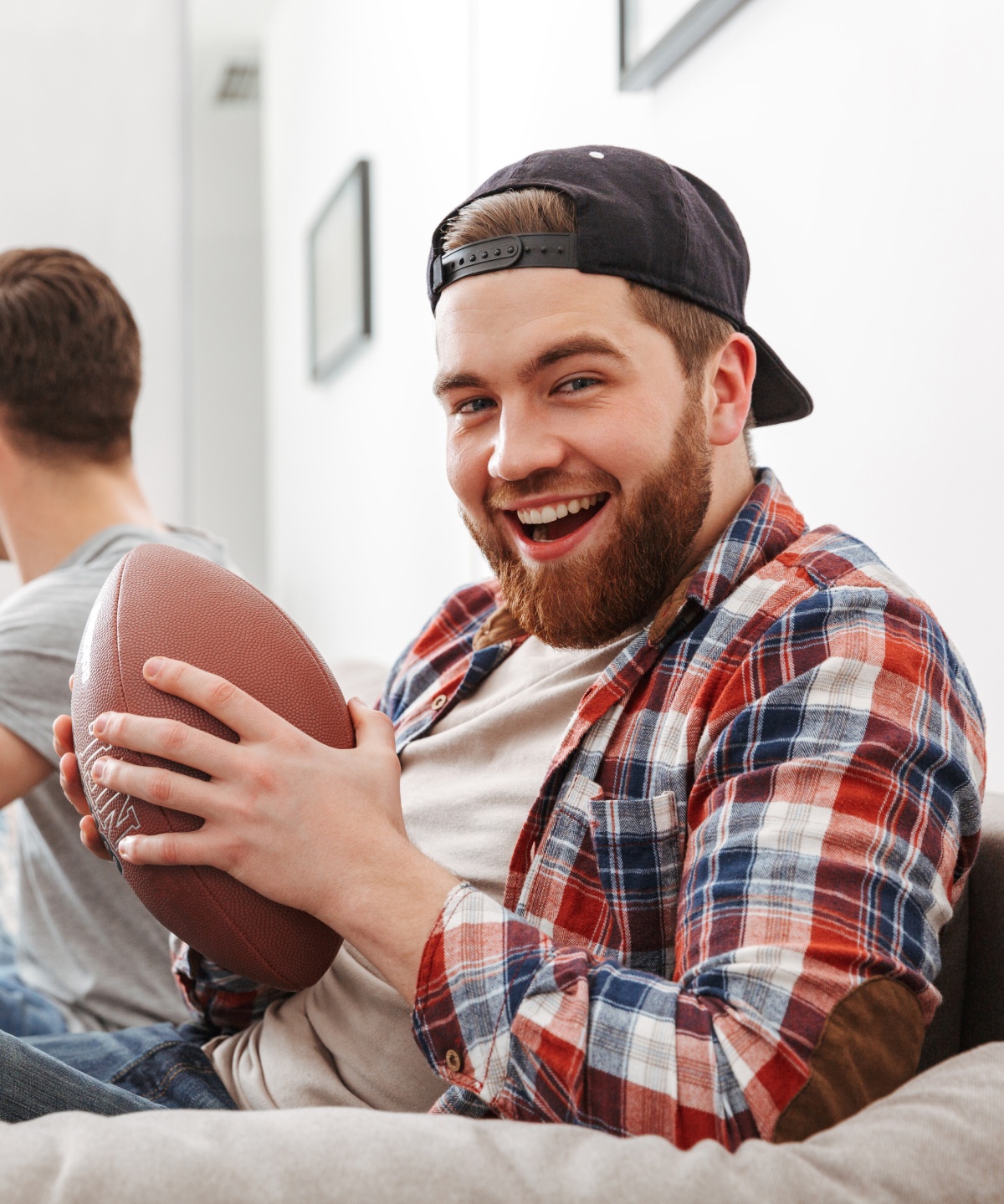 Lachender Mann mit American-Football in der Hand, sitzt auf einem Sofa, während Freunde im Hintergrund jubeln.