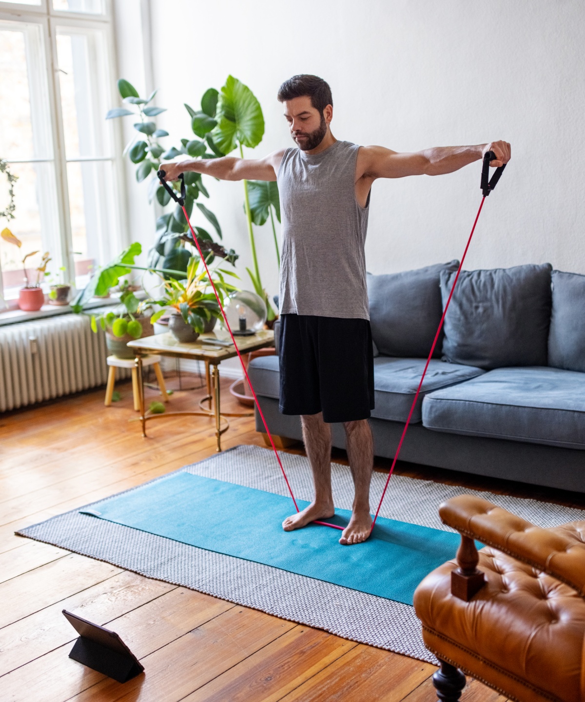 Mann trainiert mit Fitnessband auf einer Yogamatte in einem hellen modernen Wohnzimmer mit Pflanzen.
