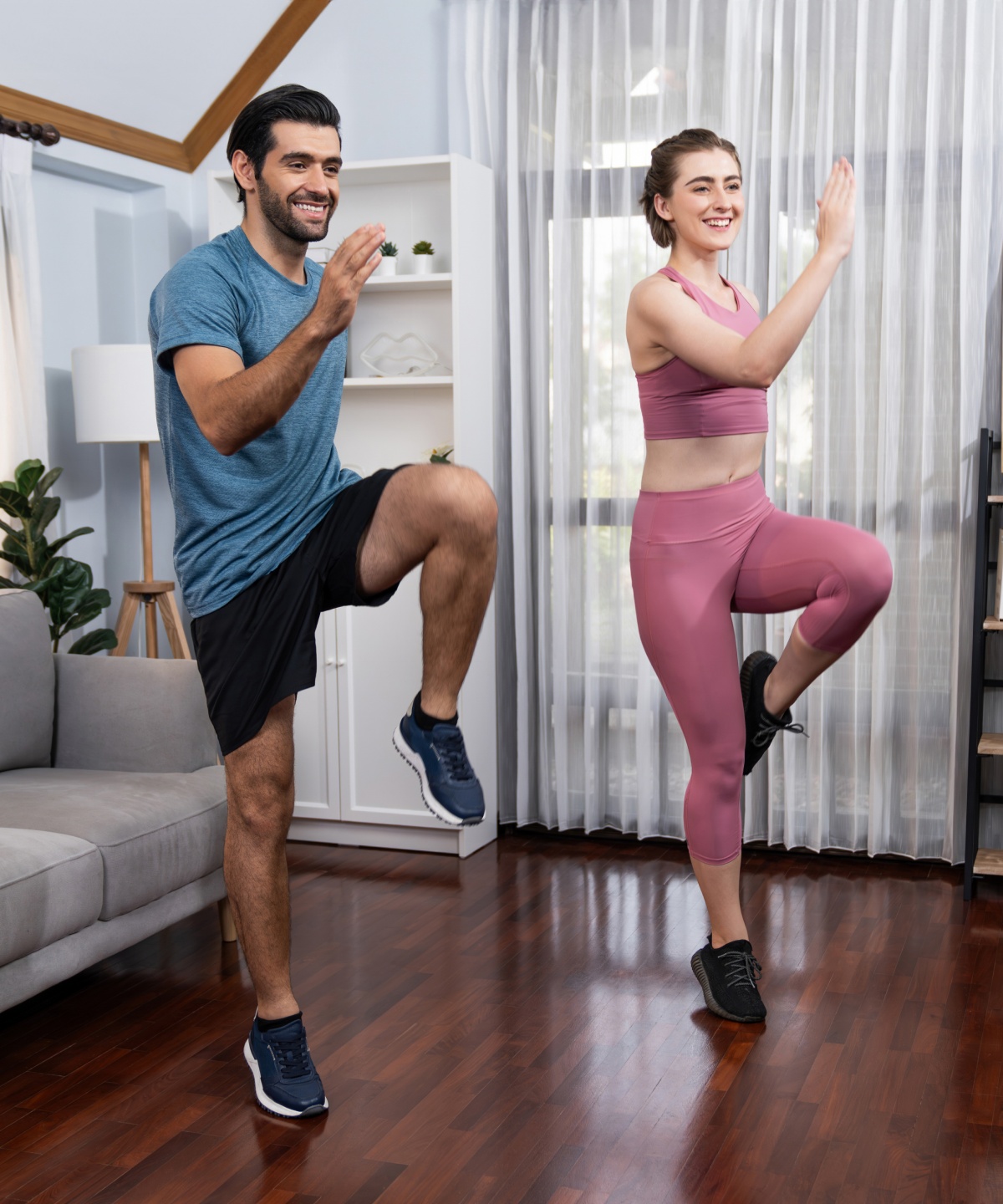 Mann und Frau trainieren gemeinsam mit Kniehebe-Übungen im Wohnzimmer zu Hause vor einem Sofa.