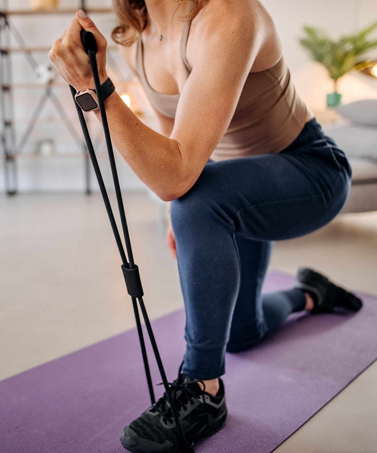 Frau kniet auf einer lila Yogamatte und trainiert mit einem Fitnessband in einem großen Wohnzimmer zu Hause.
