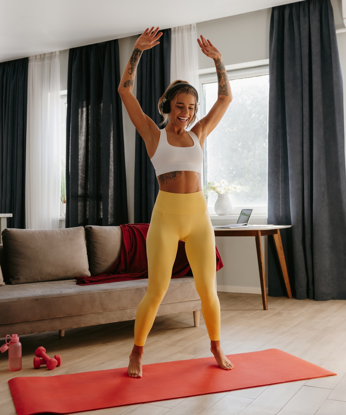 Frau in Sportkleidung und mit Kopfhörern macht stehend Übungen auf einer roten Matte in einem Wohnzimmer mit Sofa und Küchenzeile.