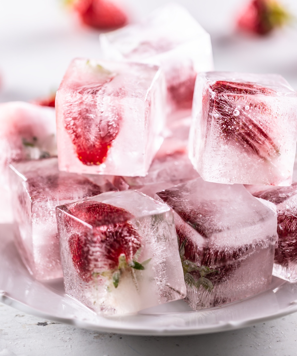 Eiswürfel mit eingefrorenen Erdbeeren auf einem weißen Teller, daneben frische Erdbeeren.