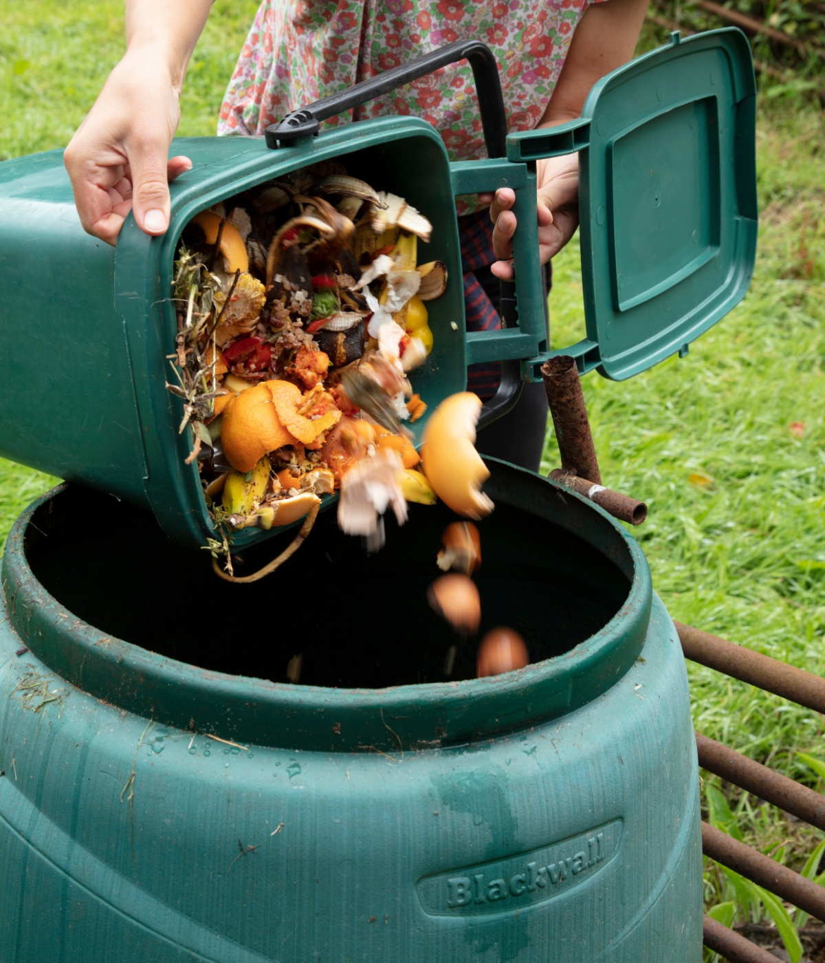 Person in geblümter Kleidung schüttet Kompost aus einem Abfalleier in eine Tonne, im Hintergrund Rasen.