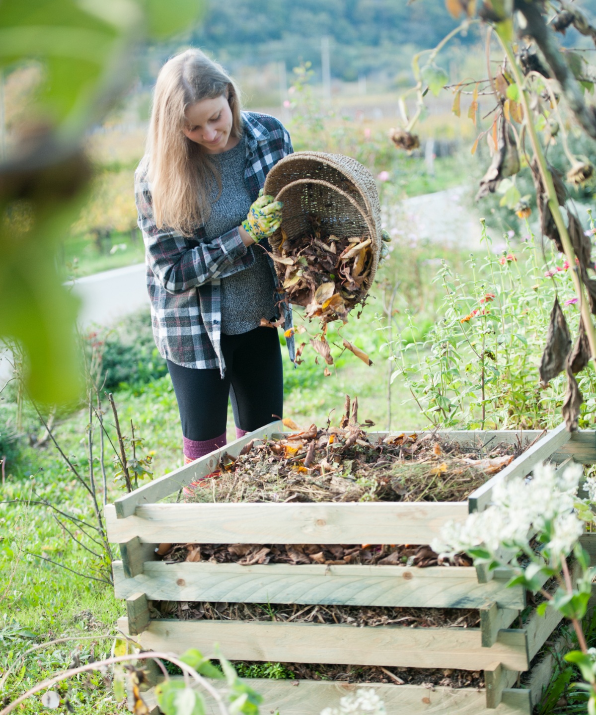 Frau im Grünen leert Gartenabfälle aus einem Korbgefäß in einen Komposter aus Holz.
