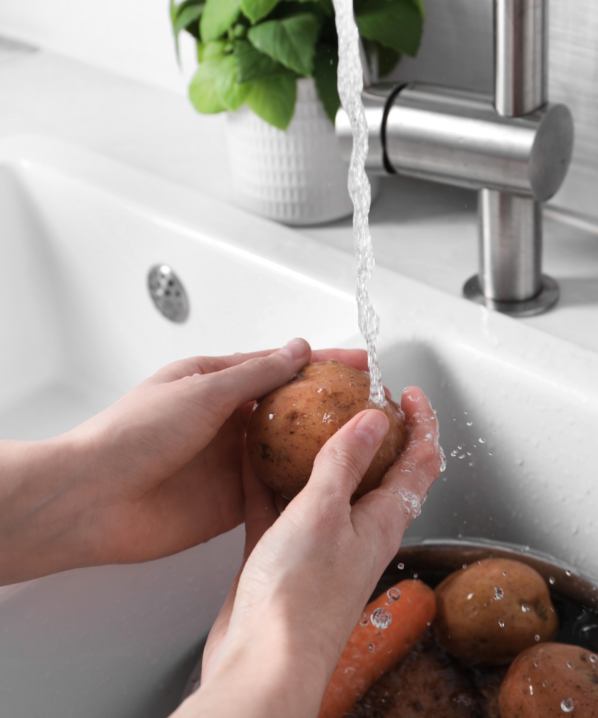 Jemand wäscht Kartoffeln unter fließendem Wasser in weißem Spülbecken ab.