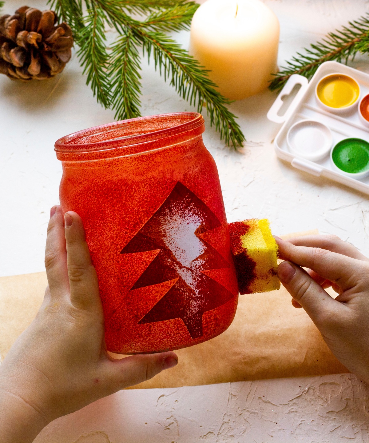 Eine Person betupft ein altes Vorratsglas mit roter Farbe und Tannenmotiv, dahinter ein Farbkasten, Tannenzweige und eine Kerze.