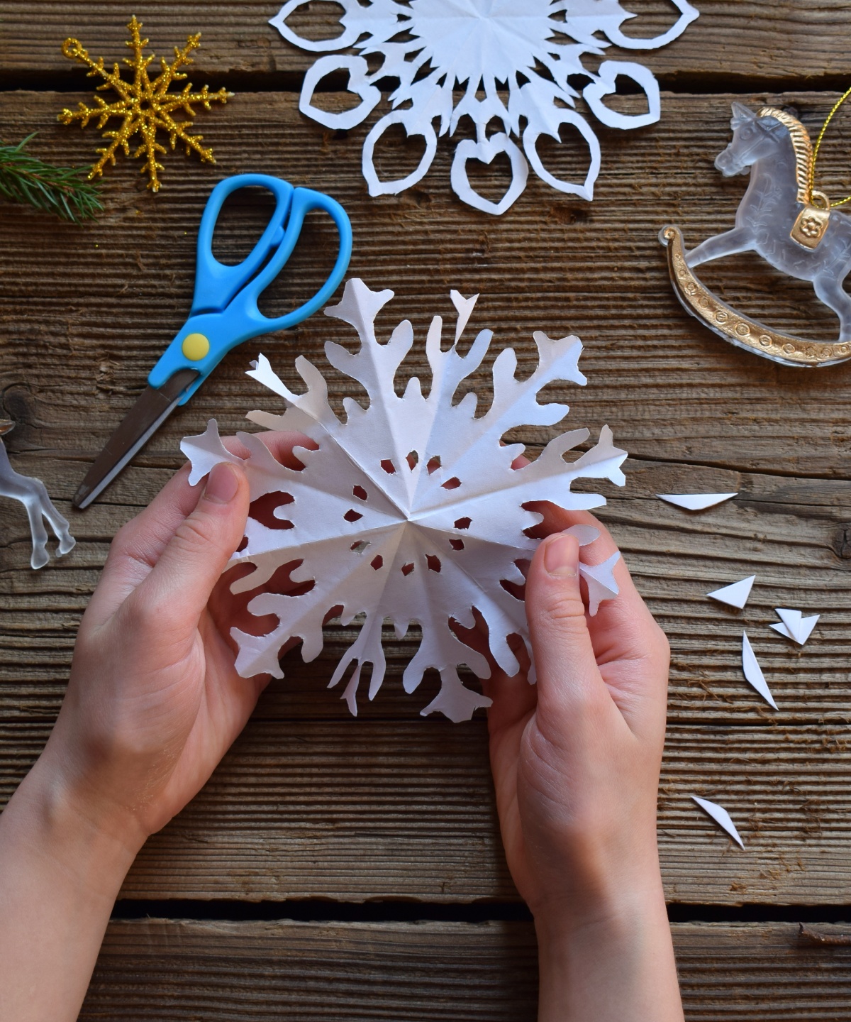 Hände basteln Schneeflocken aus weißem Papier, umgeben von Tannenzweigen und goldenen Weihnachtsdekorationen.
