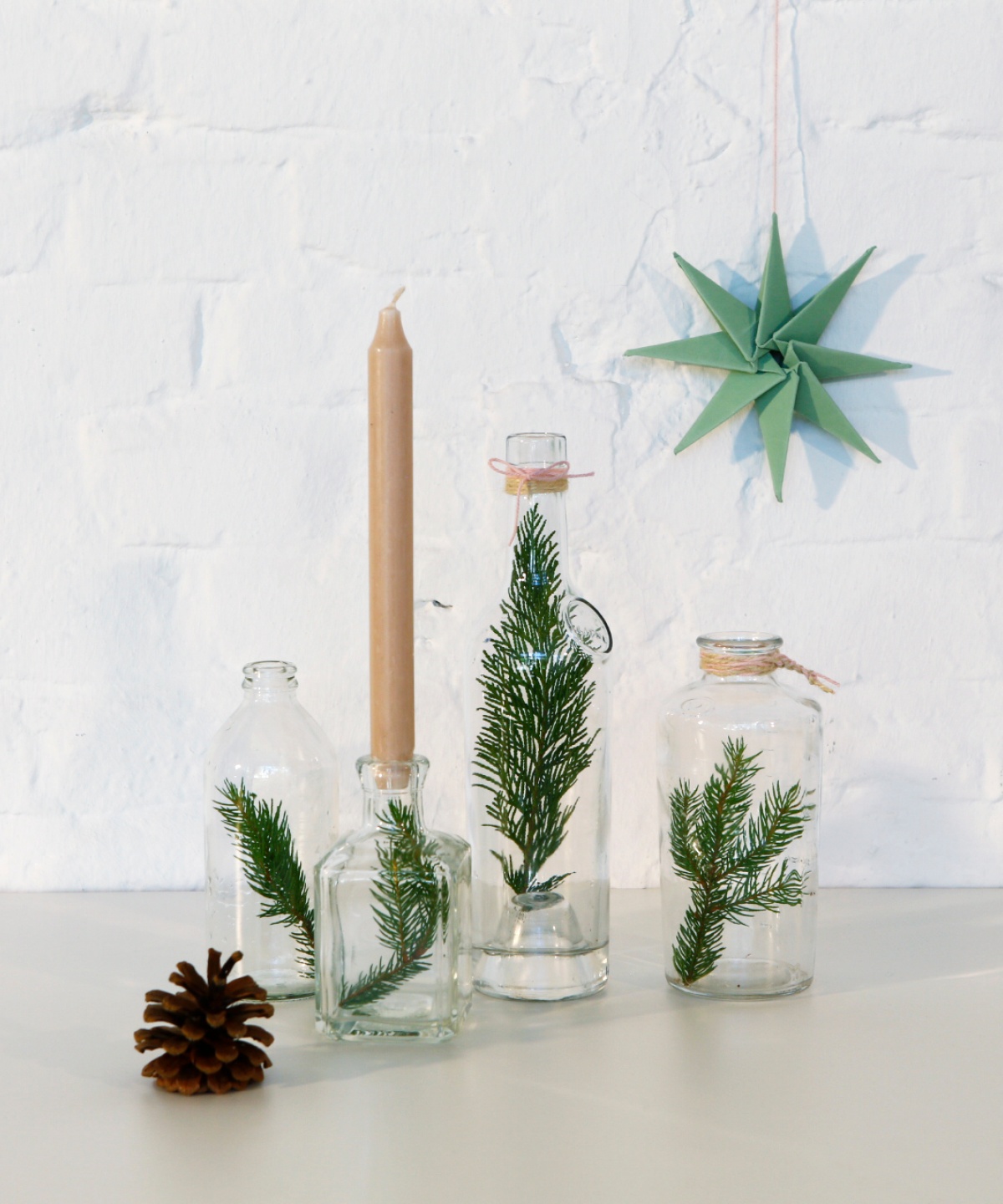 Minimalistische Weihnachtsdekoration mit Tannenzweigen in Glasflaschen, Kerze und gefaltetem Papierstern.