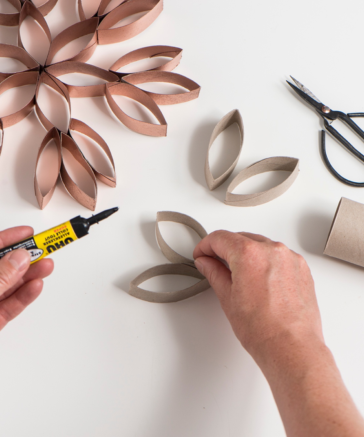 Hände basteln Schneeflocken-Deko aus Toilettenpapierrollen und Kleber, Schere im Hintergrund.