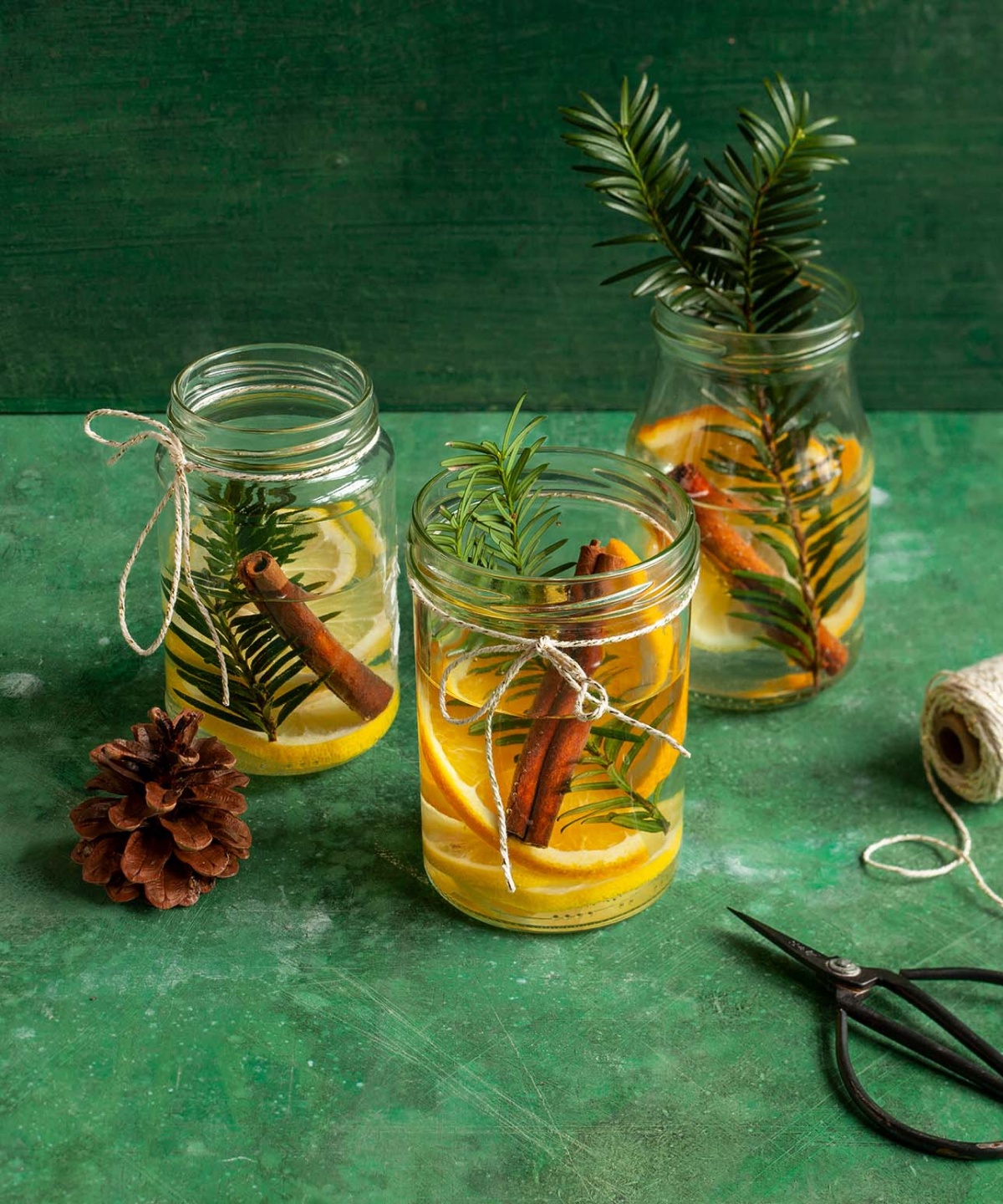 Einmachgläser mit Orangen, Zimtstangen und Tannenzweigen als festliche Tischdeko, umgeben von Garn und Tannenzapfen.