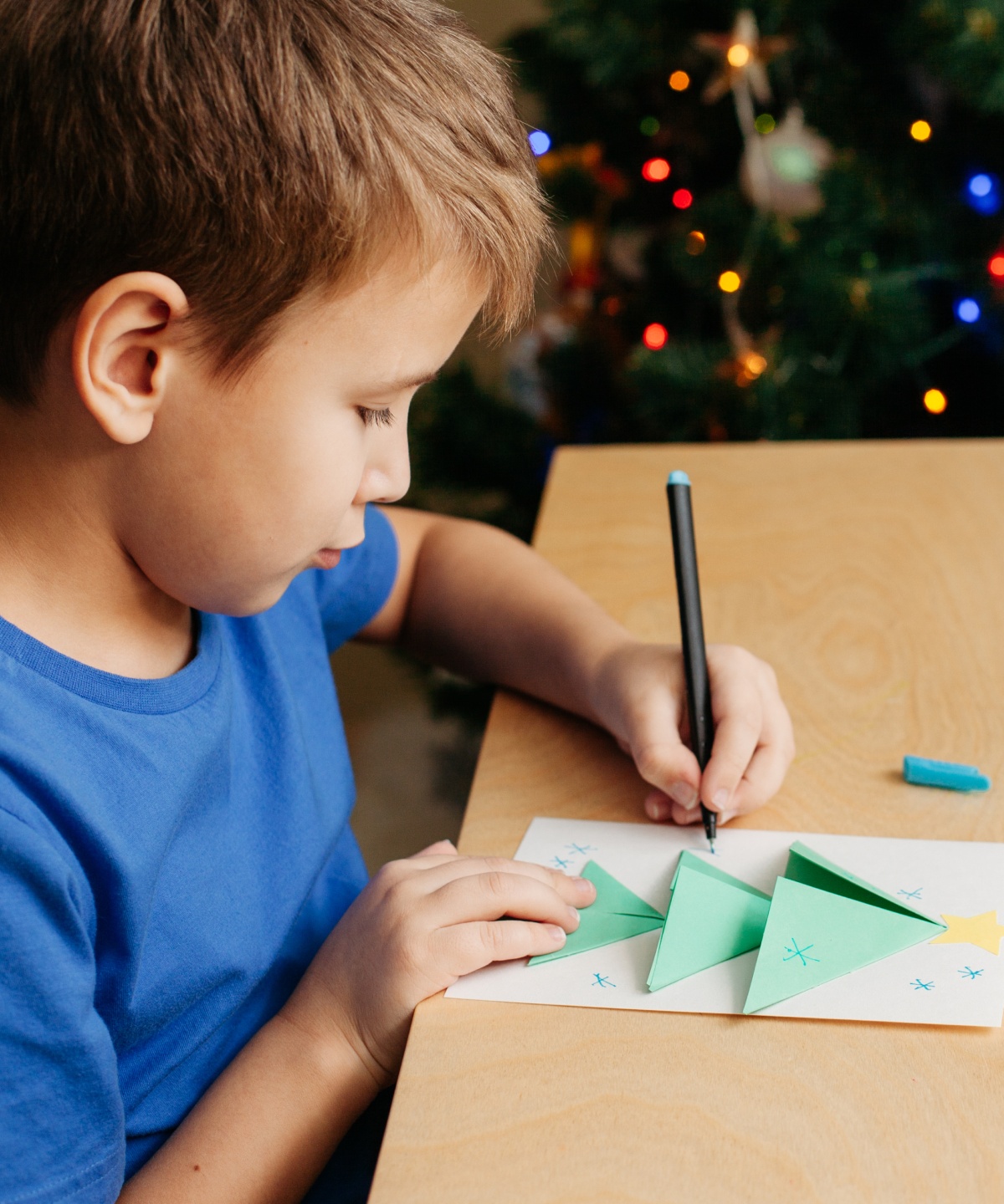 Junge bastelt Weihnachtskarte mit grünem Papier am Tisch vor geschmücktem Weihnachtsbaum.