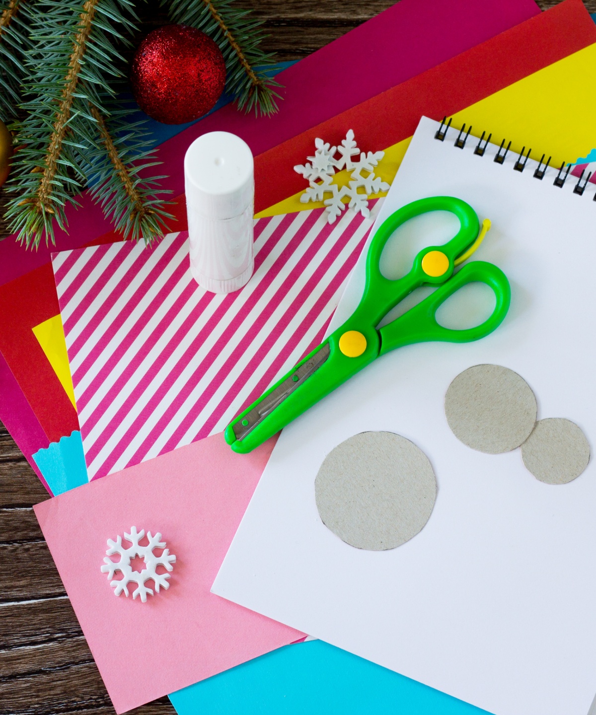 Bastelmaterialien mit Schere, Klebestift, farbigem Papier und Schneeflocken-Dekoration auf Holztisch.