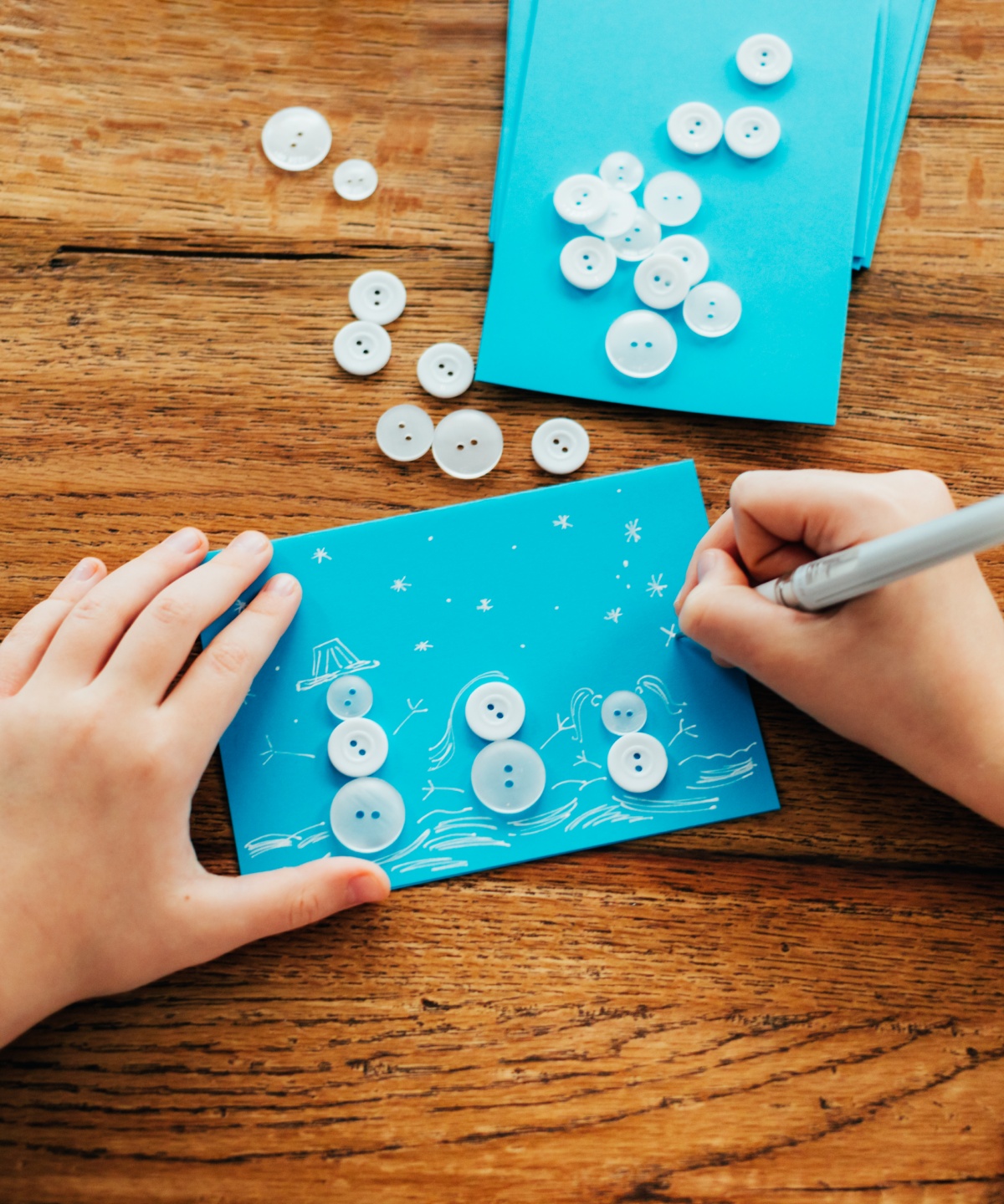 Kind bastelt Schneemannkarte aus blauen Knöpfen und zeichnet Details mit weißem Stift auf blauem Papier.