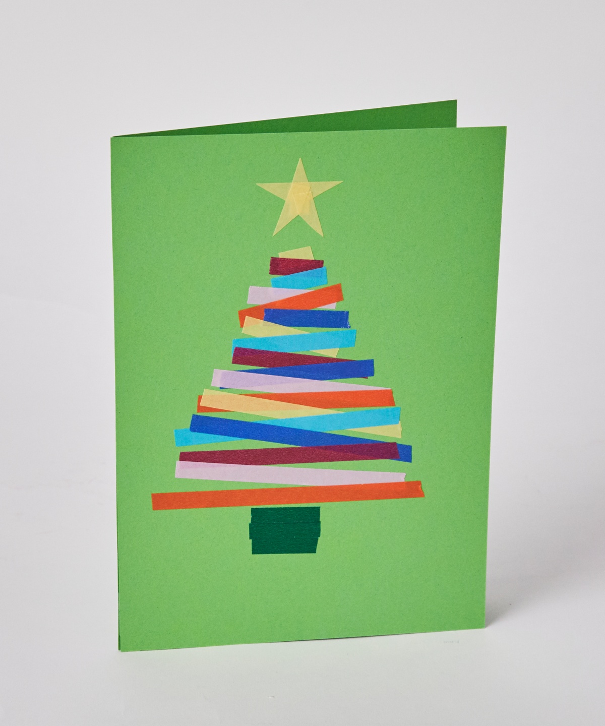 Selbstgebastelte Weihnachtskarte mit buntem Papierstreifen-Tannenbaum und gelbem Stern auf grünem Hintergrund.
