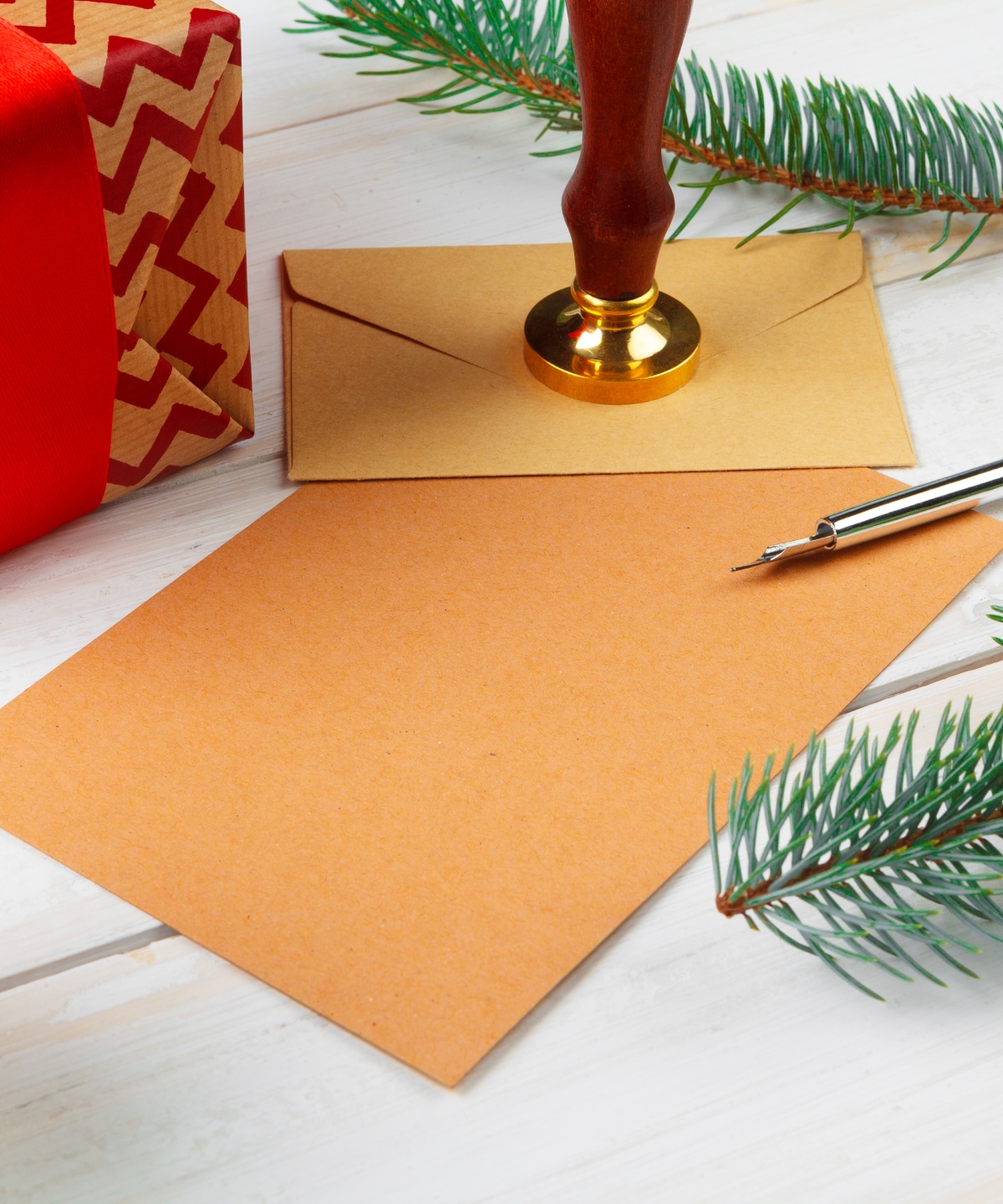 Weihnachtlich verpacktes Geschenk, Brief mit Siegelstempel, Tannenzweige und Federkiel auf hellem Holztisch.
