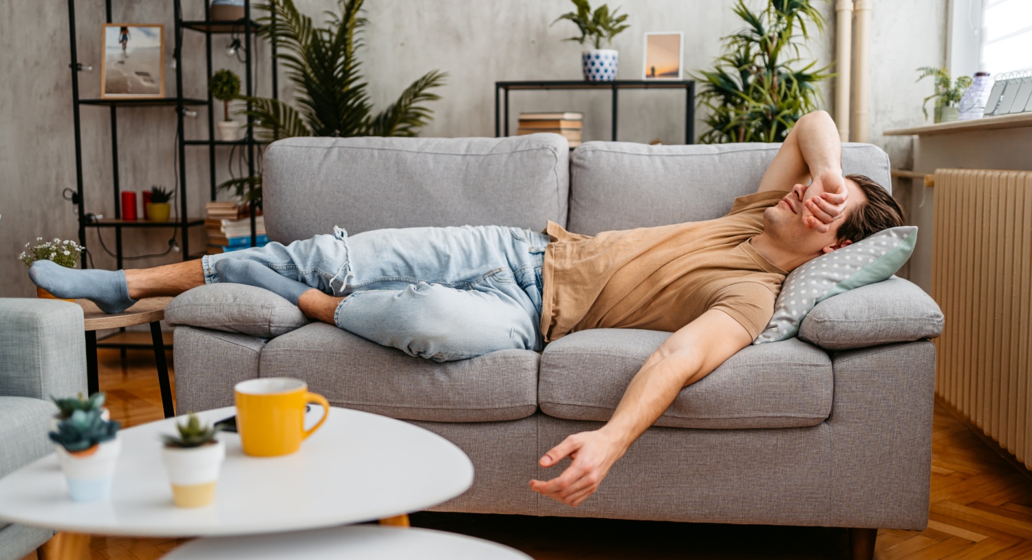 Mann liegt erschöpft auf einer grauen Couch und hält sich die Stirn.