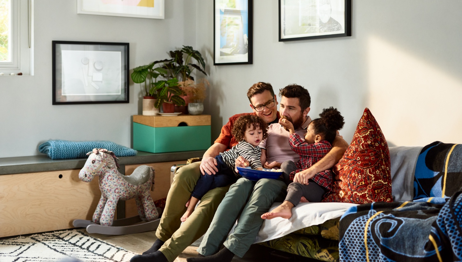 Zwei Männer sitzen mit ihren Kindern gemütlich auf dem Sofa im Wohnzimmer