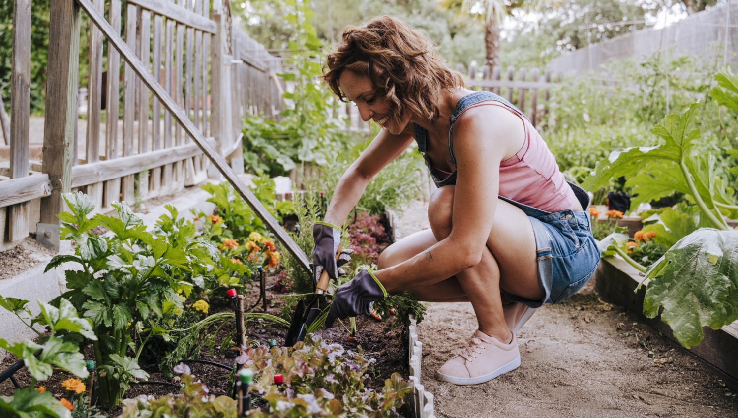 Frau hockt in kurzer Jeans-Latzhose, Tanktop und Gartenhandschuhen in einem mit Holzlatten eingezäunten Garten auf einem Weg. Links und rechts von ihr sind Beete angelegt, sie gräbt in dem linken Beet mit einer kleinen Schaufel.