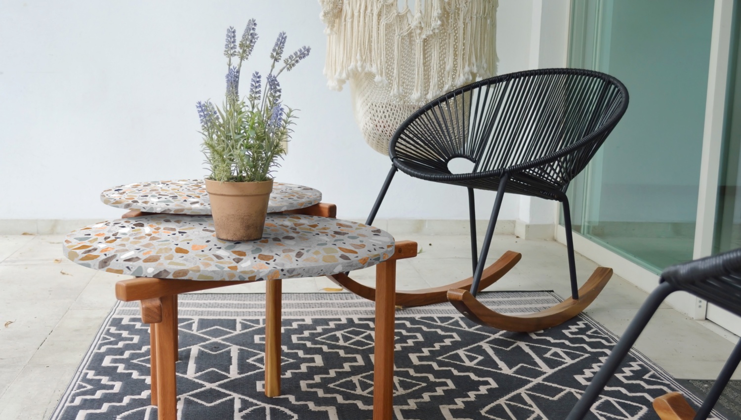 Stilvolle Terrasse mit schwarzem Schaukelstuhl, Terrazzo-Tischen, Lavendelpflanze und Makramee-Deko vor weißer Wand.