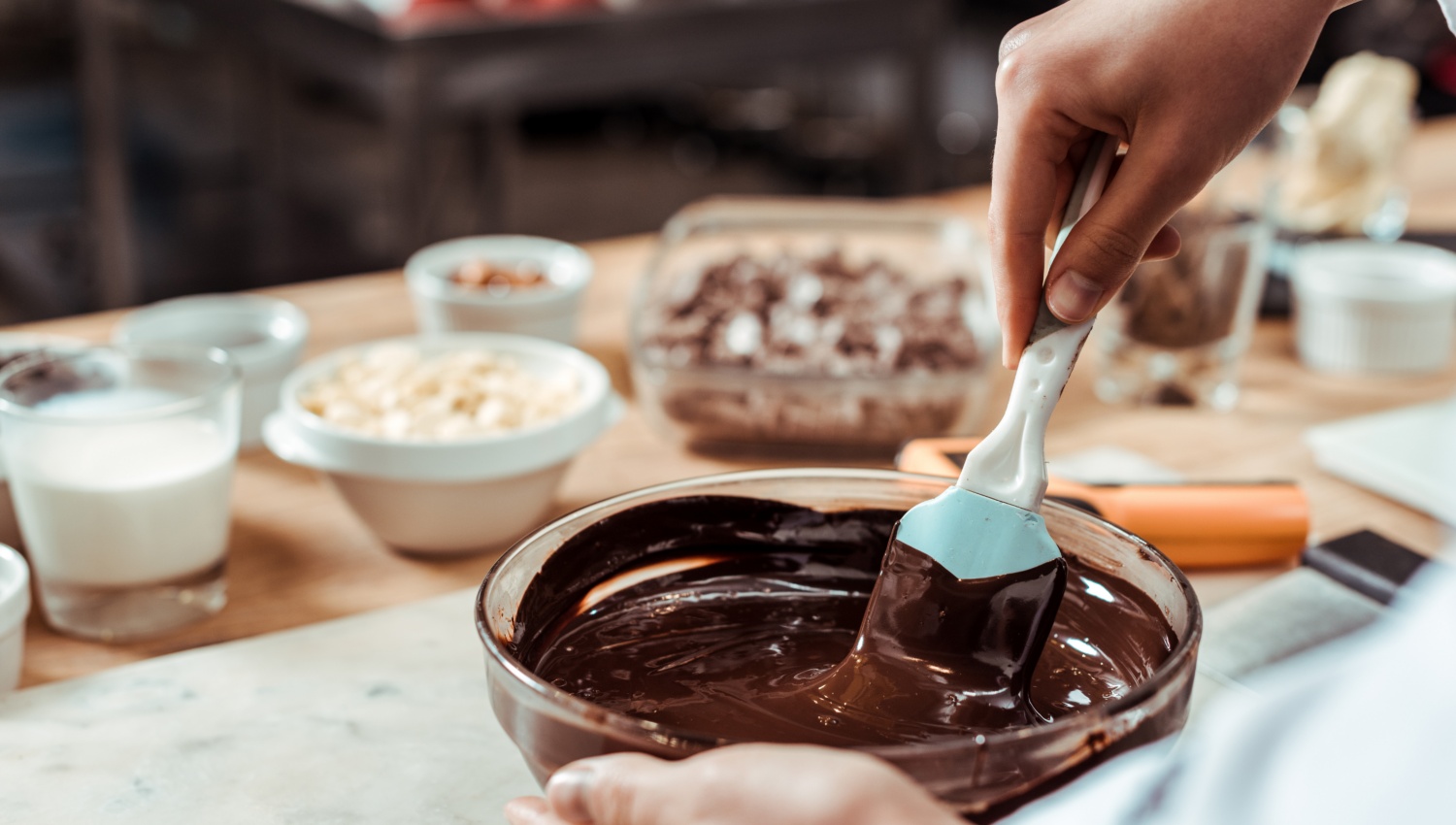 Hand rührt geschmolzene Schokolade in einer Glasschüssel. Im Hintergrund stehen Zutaten wie Milch, Nüsse und Schokostücke.