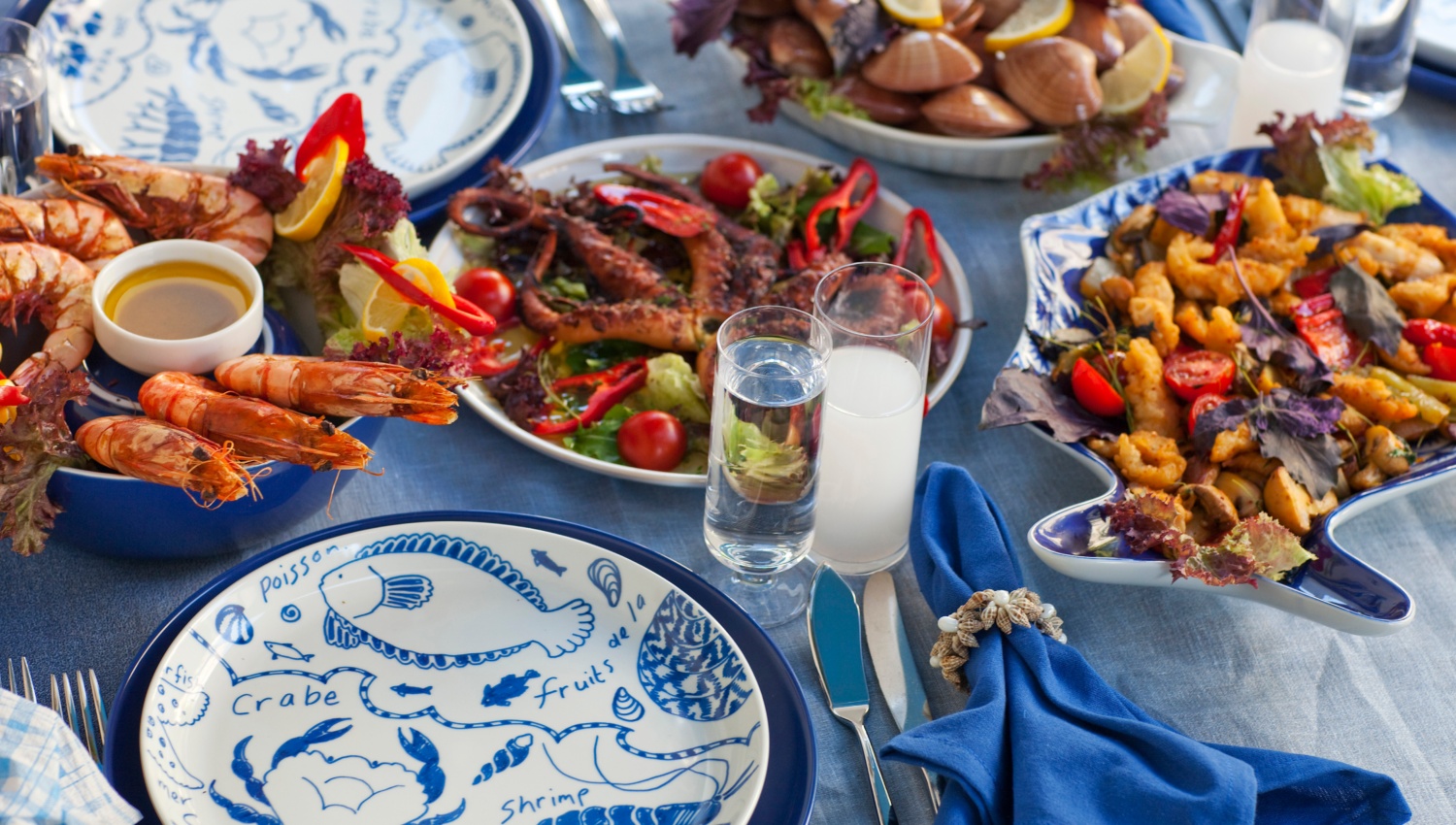 Ein üppig gefüllter Tisch mit traditionell griechischem Essen, blau-weißem Geschirr und blauen Servietten.