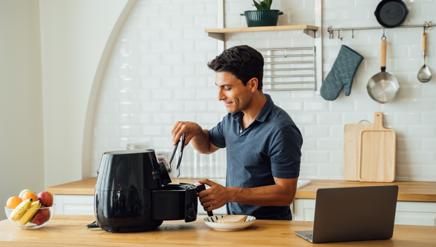 Ein Mann steht mit einer Küchenzange in der Hand vor einer geöffneten Heißluftfritteuse und schaut hinein.