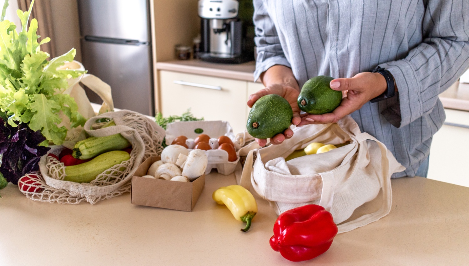 Person hält zwei Avocados über einem Tisch mit frischem Gemüse und Eiern in einer Küche.