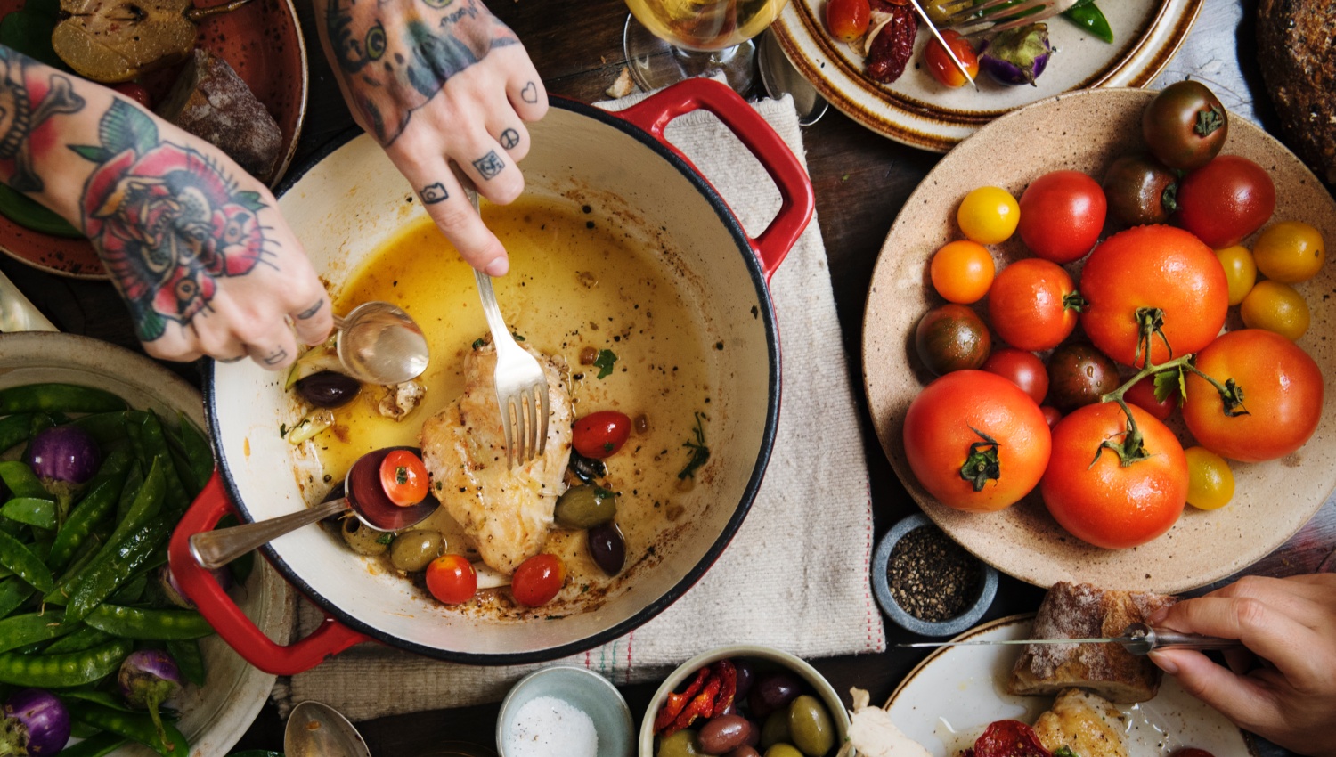 Reich gedeckter Tisch von oben, eine Frau mit tätowierten Händen holt mit einer Gabel eine gebratene Hähnchenbrust mit Tomaten aus einem Brattopf, rechts vom Topf steht eine Schale mit vielen verschiedenfarbigen Tomaten, links vom Topf steht eine Schale mit grünen Bohnen.