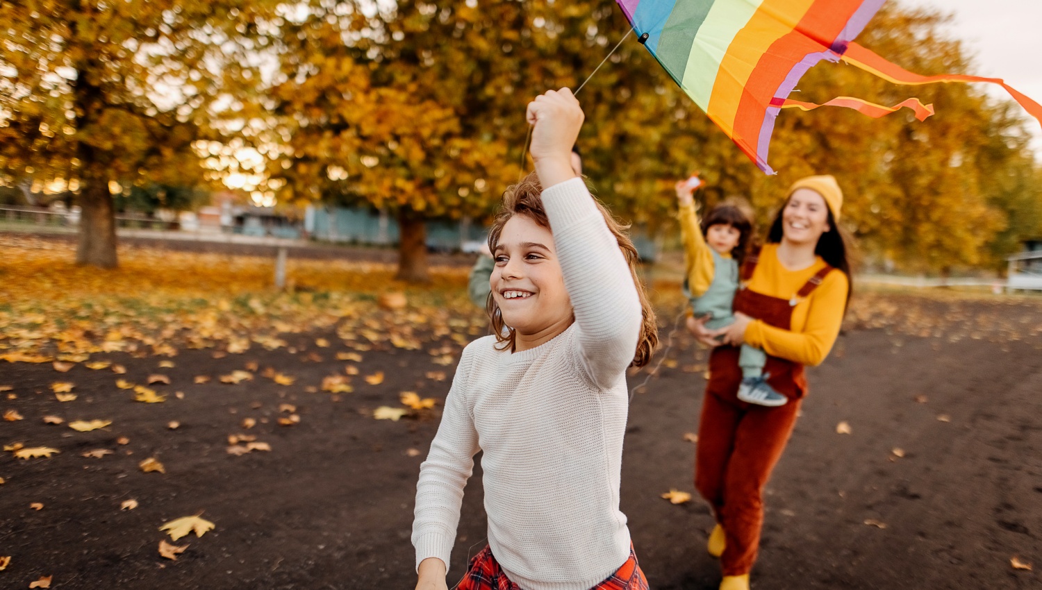Kind lässt einen selbst gebastelten, bunten Drachen im Herbstpark fliegen, während eine Mutter mit Kleinkind zuschaut.