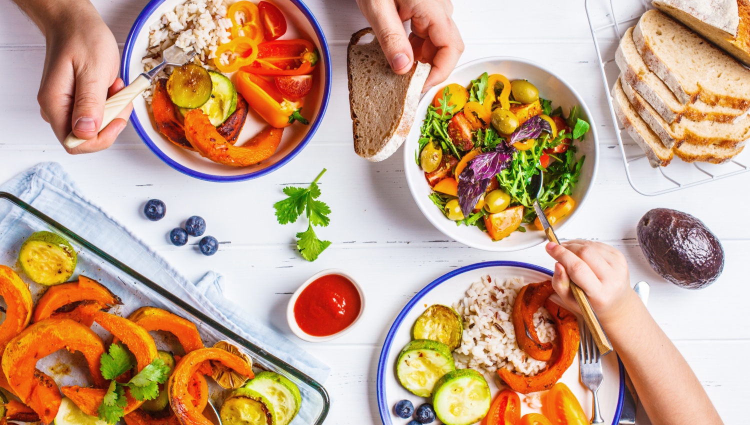 Ein reich gedeckter Tisch mit Tellern und Schüsseln voller Gemüse. Kinder- und Erwachsenenhände bedienen sich.
