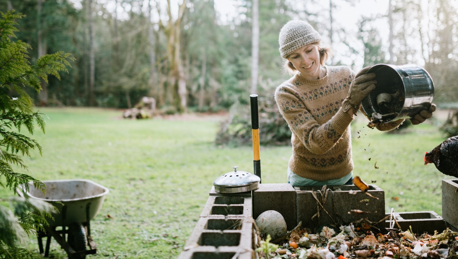 Frau in Mütze, Pullover und Gartenhandschuhen leert Abfalleimer in einen Komposter. Rechts neben ihr ein Huhn, links eine Schubkarre, im Hintergrund ein großer Garten mit hoch gewachsenen Bäumen.