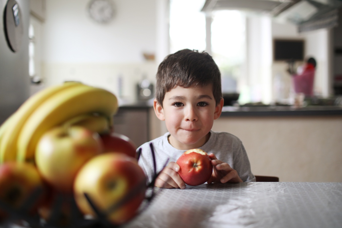Junge isst einen Apfel am Küchentisch, vor einem Obstkorb.