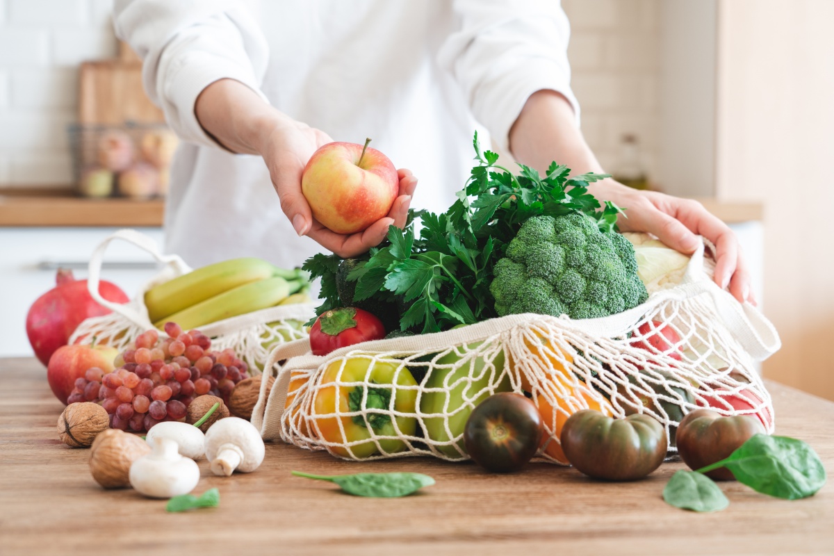 Person holt einen Apfel aus einer großen Netz-Einkaufstasche, die gefüllt ist mit viel frischem Gemüse.