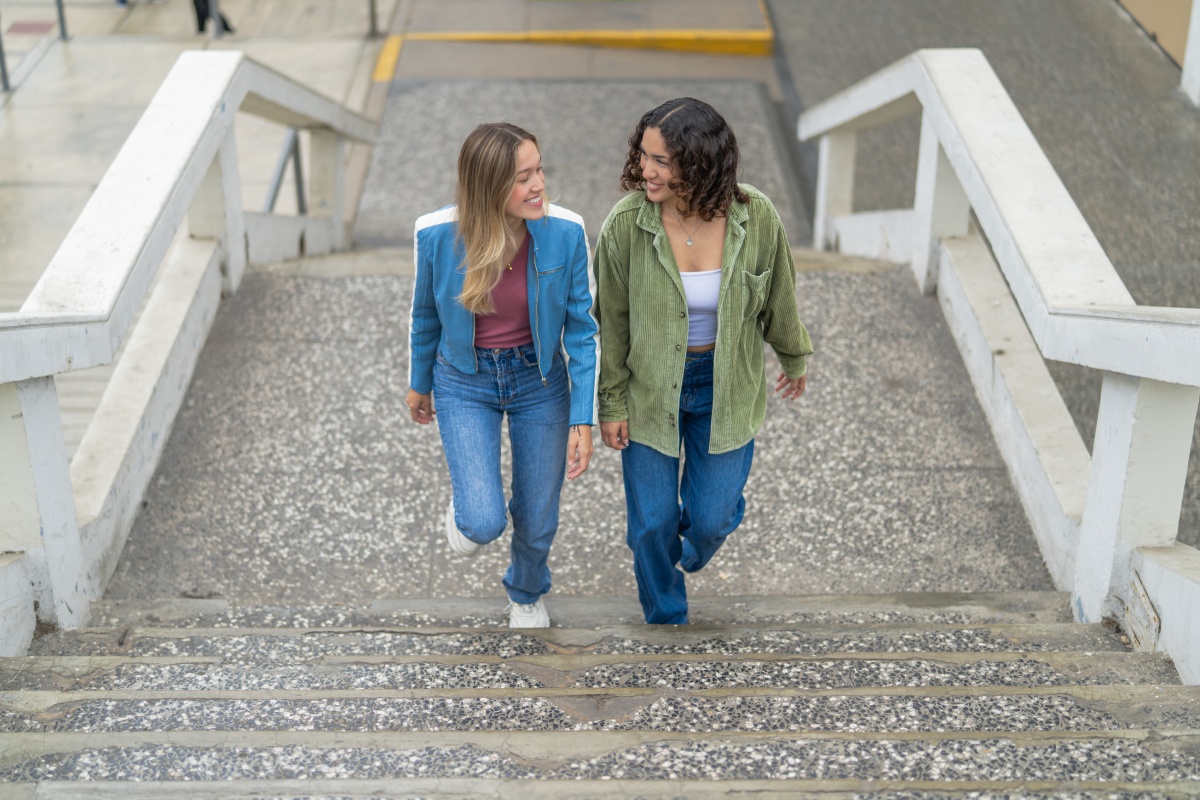 Zwei junge Frauen in lässiger Kleidung gehen lächelnd eine Treppe im Freien hinauf.