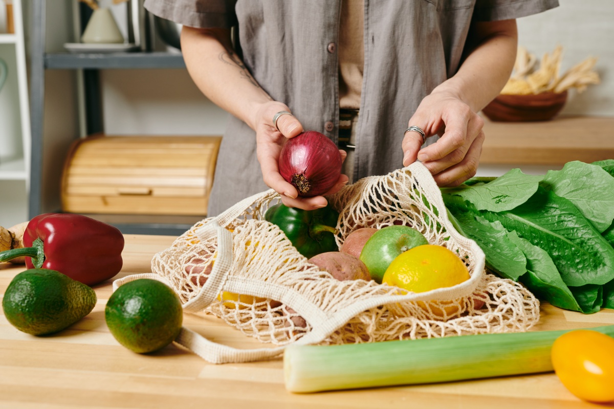 Person packt frisches Gemüse und Obst, darunter rote Zwiebel, Paprika und Avocados, aus einem Netzbeutel aus.