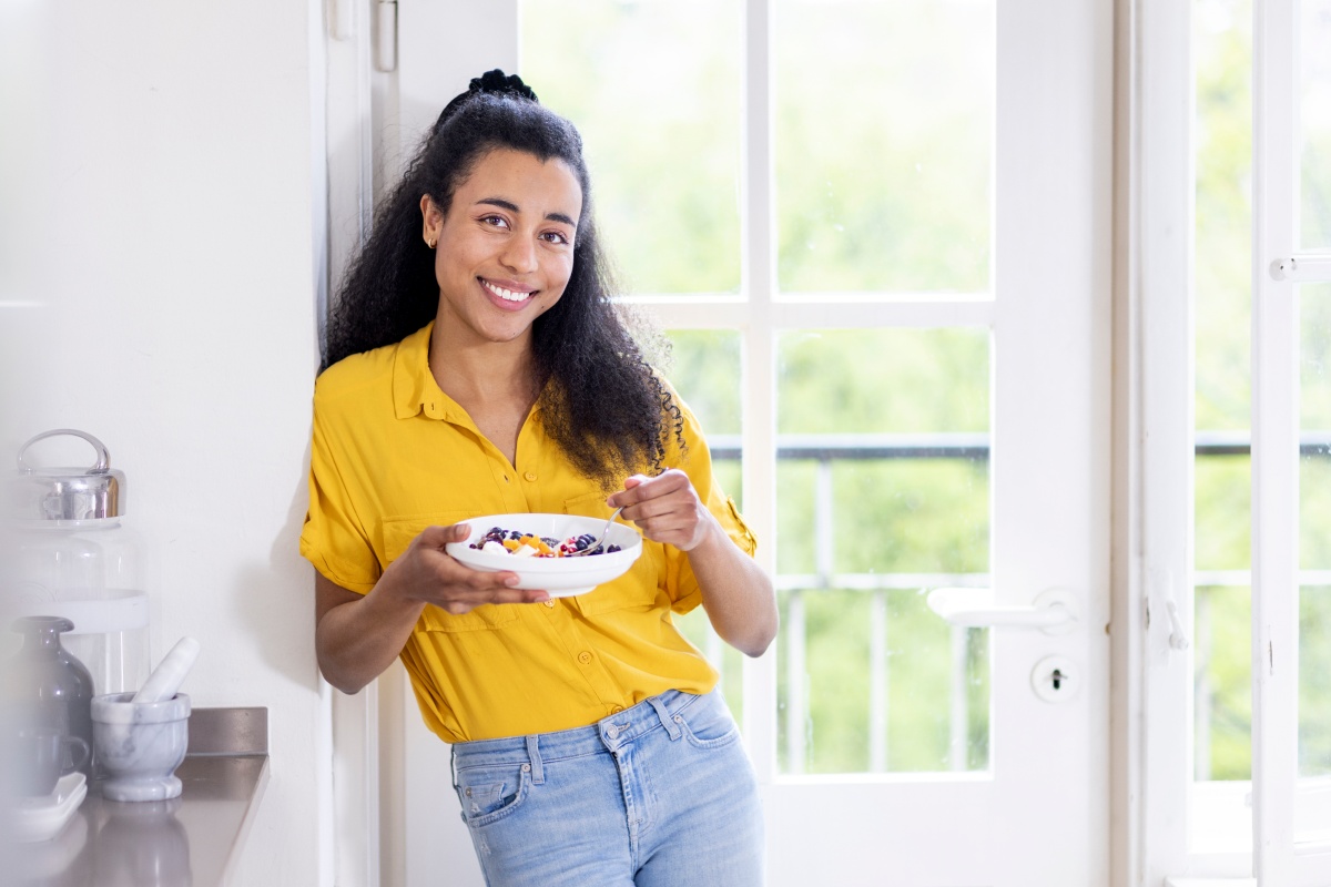 Lächelnde Frau in gelber Bluse steht mit einer Frühstücksschale vor einem hellen Fenster.