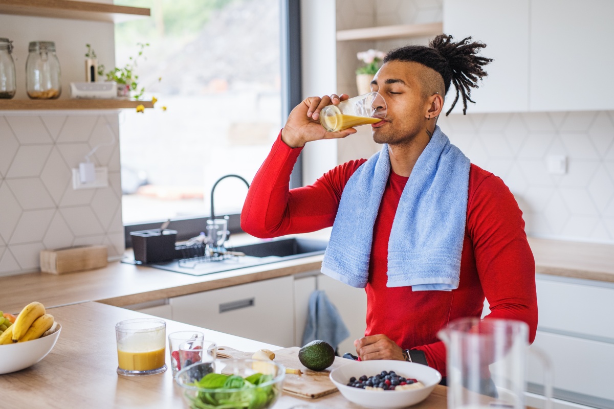 Sportlicher Mann in rotem Shirt trinkt Saft in einer modernen Küche, ein Handtuch um den Hals, Obst und Gemüse auf der Theke.