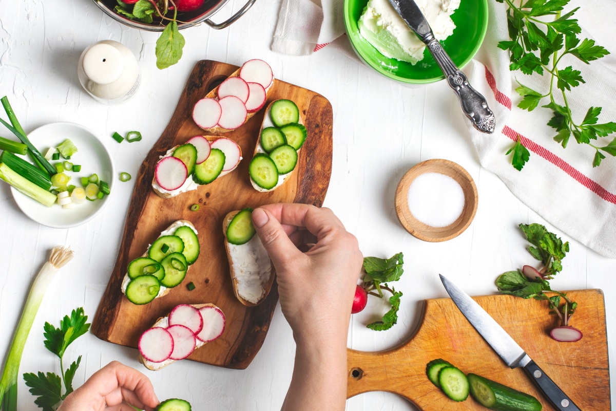 Frische Brot-Scheiben werden mit Gurken- und Radieschenscheiben auf einem Holzbrett belegt.