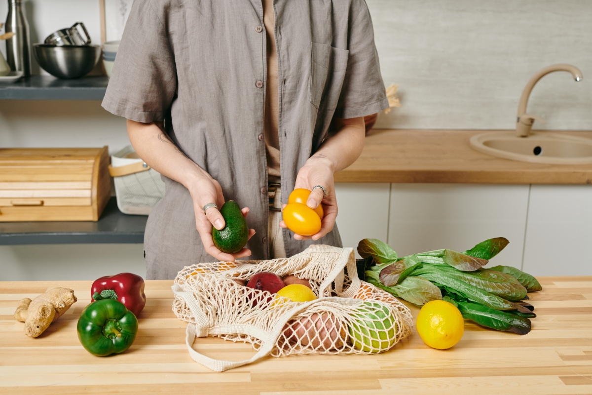 Person in einer Küche nimmt eine Avocado und gelbe Tomaten aus dem Netzbeutel, der mit frischem Gemüse gefüllt ist.