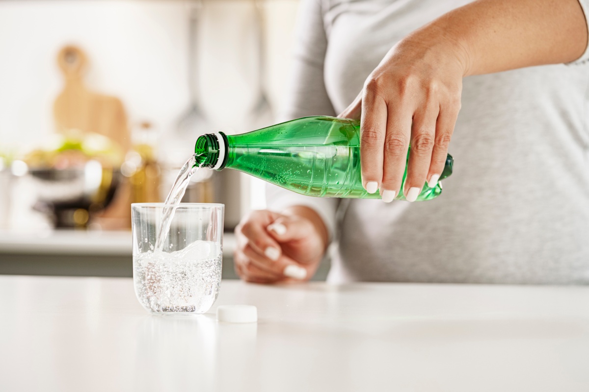 Person gießt Mineralwasser aus einer grünen PET-Flasche in ein Glas, das auf einer Küchenarbeitsplatte steht.