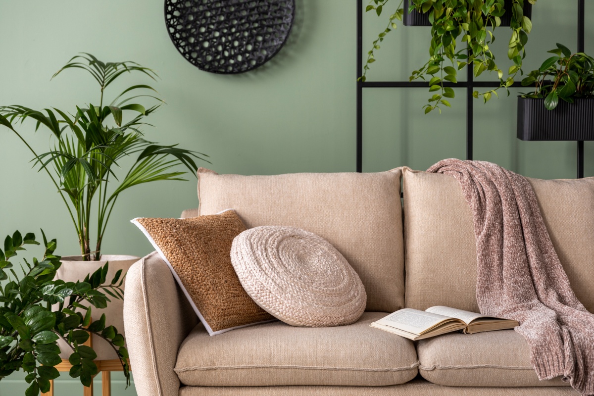 Ein gemütliches Sofa dekoriert mit Decken und Kissen steht in einem Raum. Im Hintergrund ein warmer grüner Farbton an der Wand und grüne Pflanzen