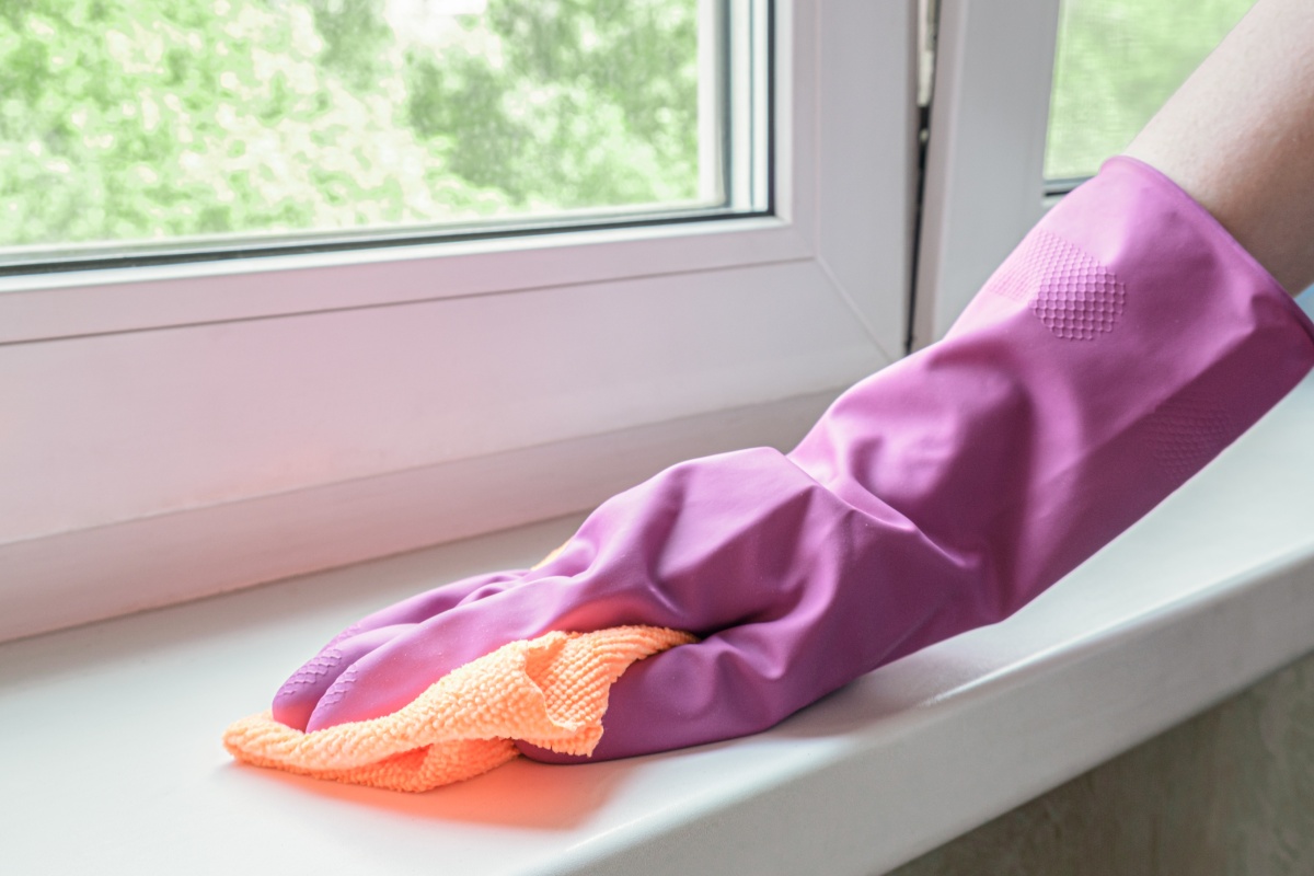 Eine Person, die Putzhandschuhe trägt, wischt Schmutz mit einem Lappen von der Fensterbank.