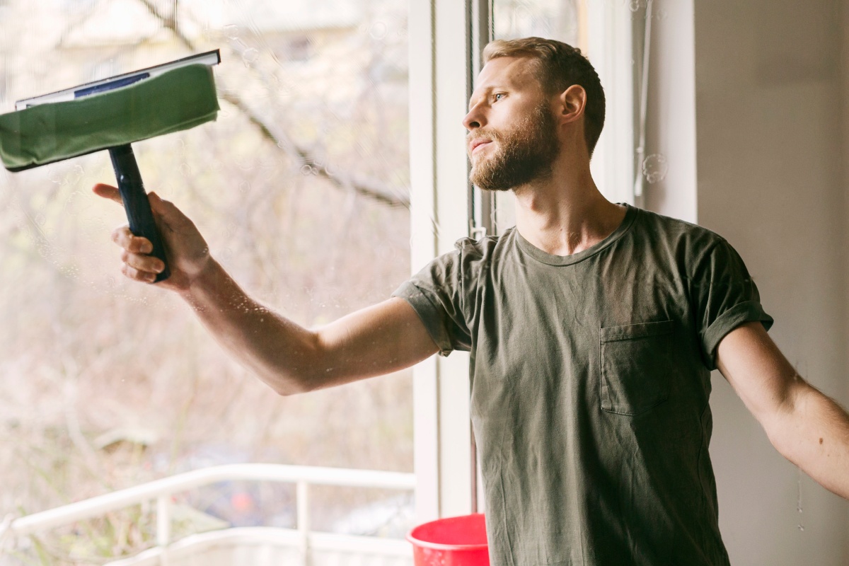 Ein Mann putzt sein Balkonfenster mit einem Abzieher. Neben ihm steht ein Eimer mit Putzwasser.