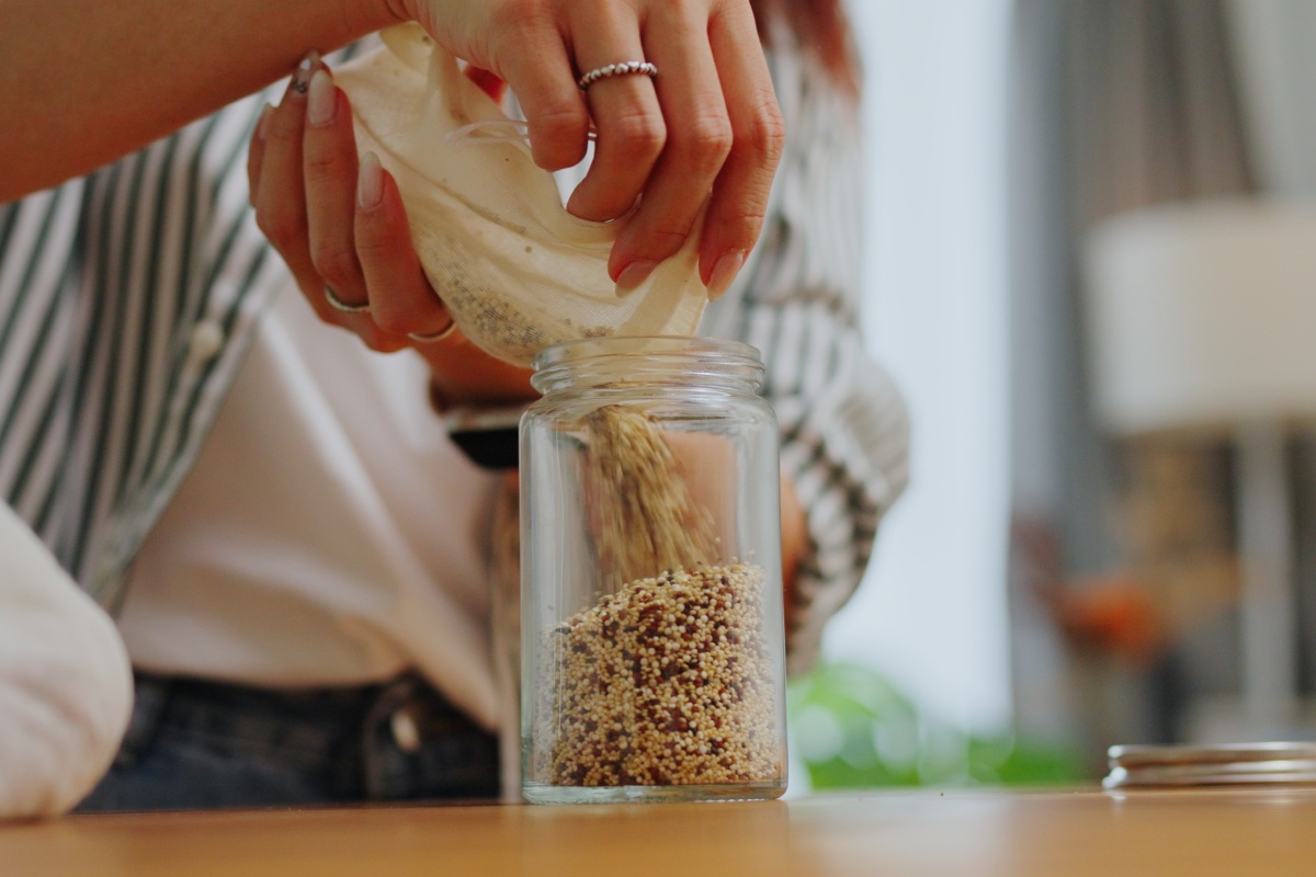 Nahaufnahme von Händen, die Quinoa aus einem Beutel in ein Glasgefäß füllen.