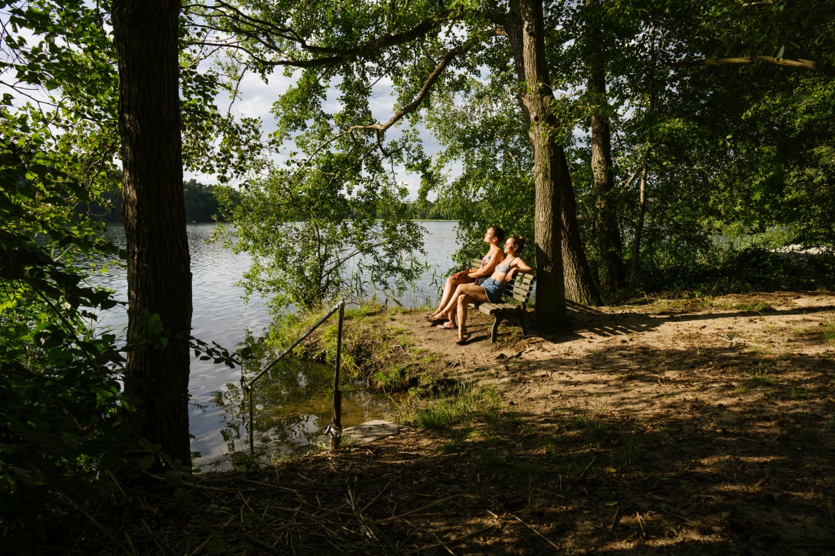 Zwei Frauen sitzen auf eine Holzbank an einem Waldsee, den Blick in die Sonne gerichtet.