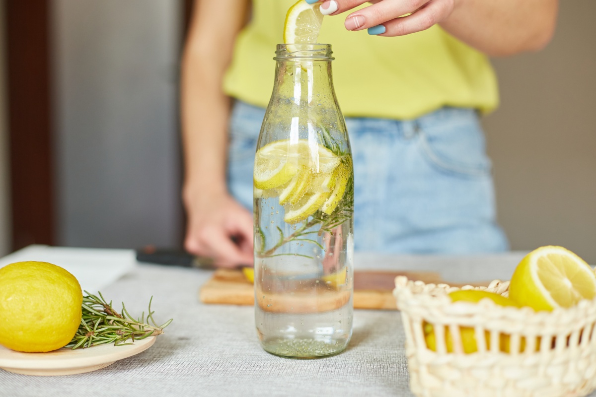 Person fügt Zitronenscheibe in eine Flasche Wasser mit Rosmarin, daneben Zitronen und ein Korb.