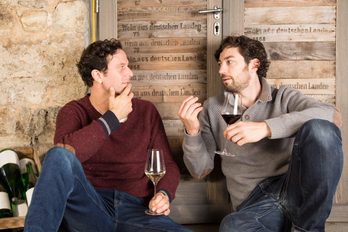Zwei Winzer unterhalten sich über die Qualität von Wein. Dabei sitzen sie vor einer Holztür mit einem Glas Wein in der Hand.