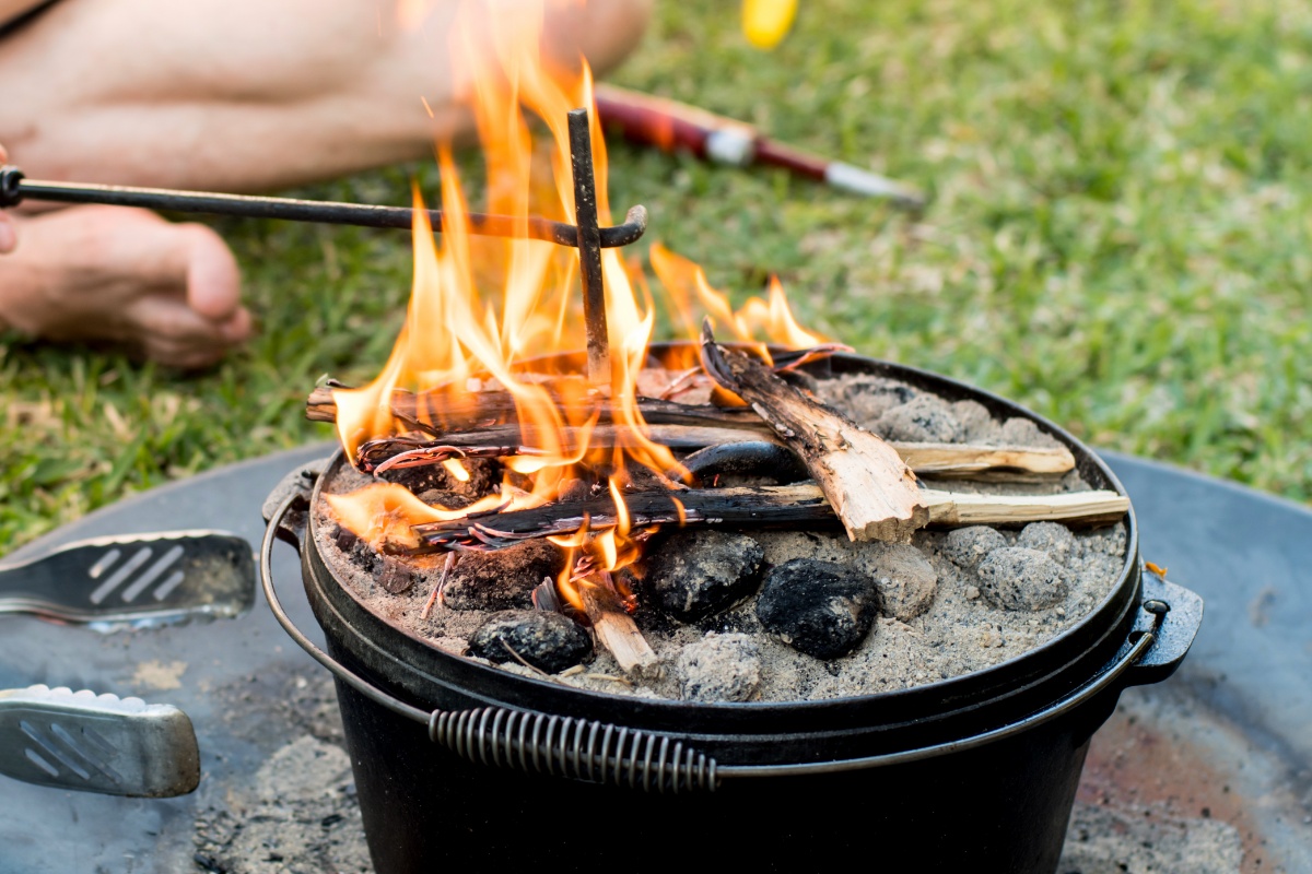 Im Gras stehender Dutch Oven, in dem ein kleines Feuer aus Kohle und Holz brennt, das Bein einer Person im Schneidersitz ist zu erkennen, die mit einem Stab das Feuer schürt.