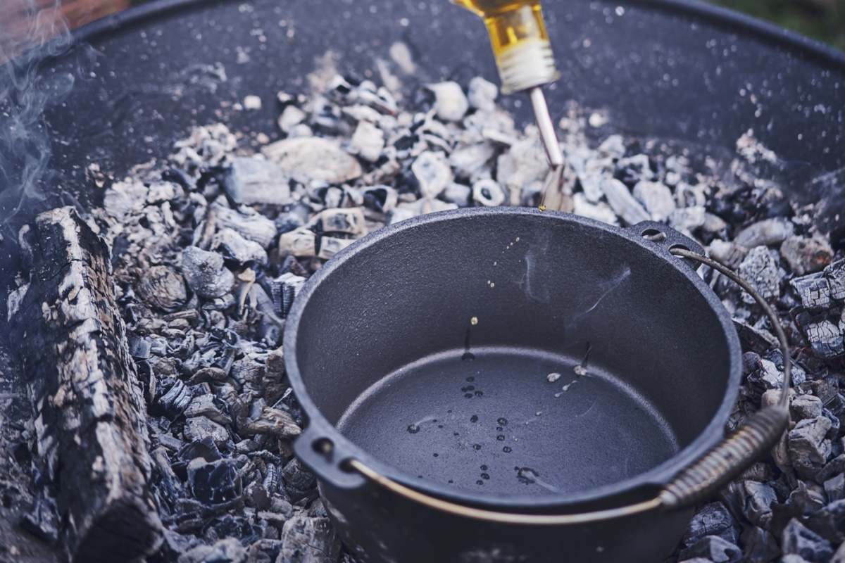 Person gießt Öl aus einer Flasche in einen Dutch Oven-Topf, der in einer mit Kohle erhitzten Feuerschale steht. Im Hintergrund unscharf Garten und Grillzubehör.